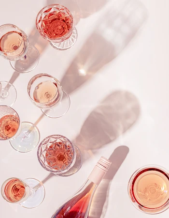 A top-down view shows a cluster of wine glasses and a bottle, with pink and rose tones casting soft shadows on a pale surface.